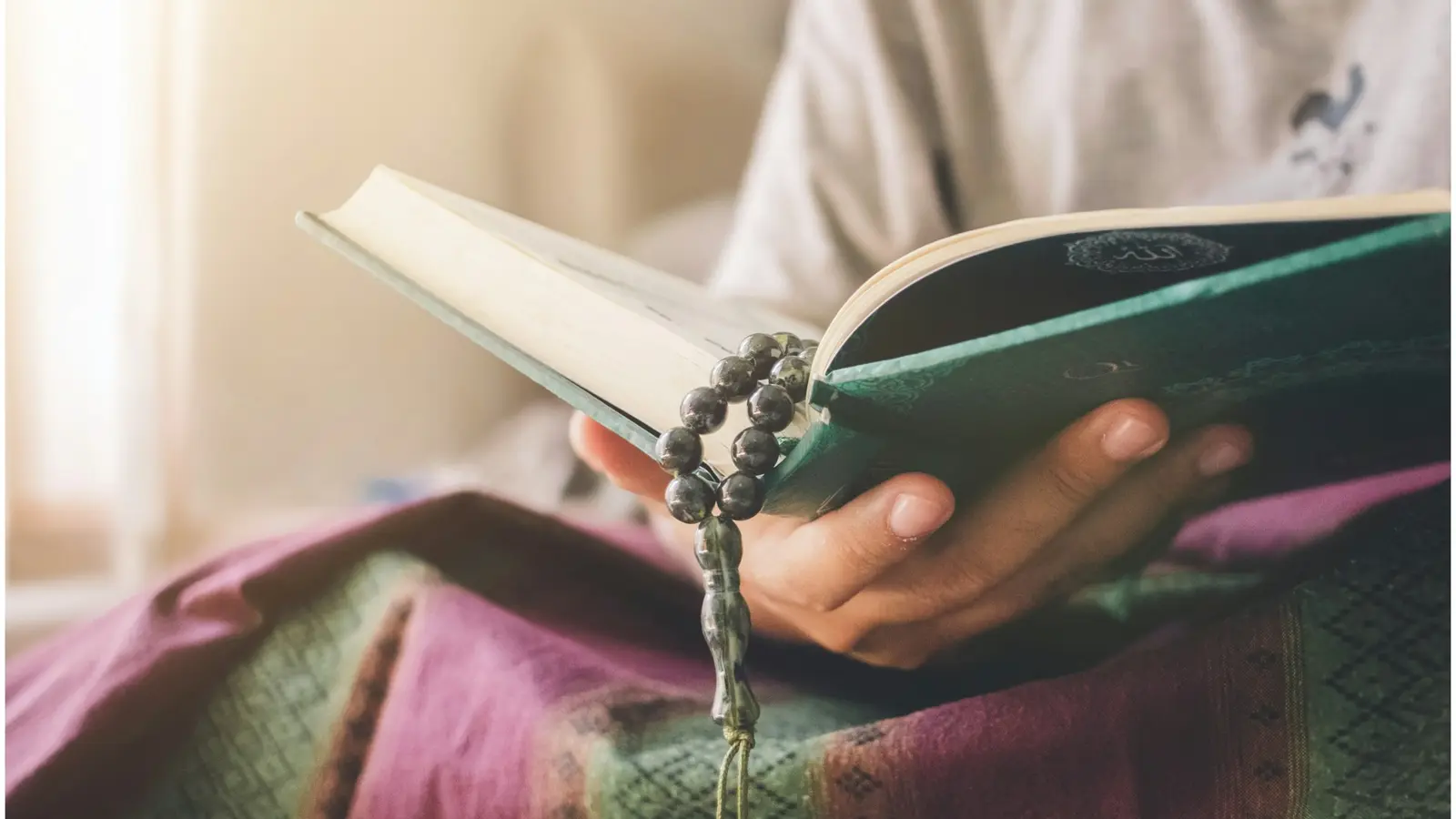 Hands holding an open Qur’an with a string of prayer beads, resting on a patterned cloth.