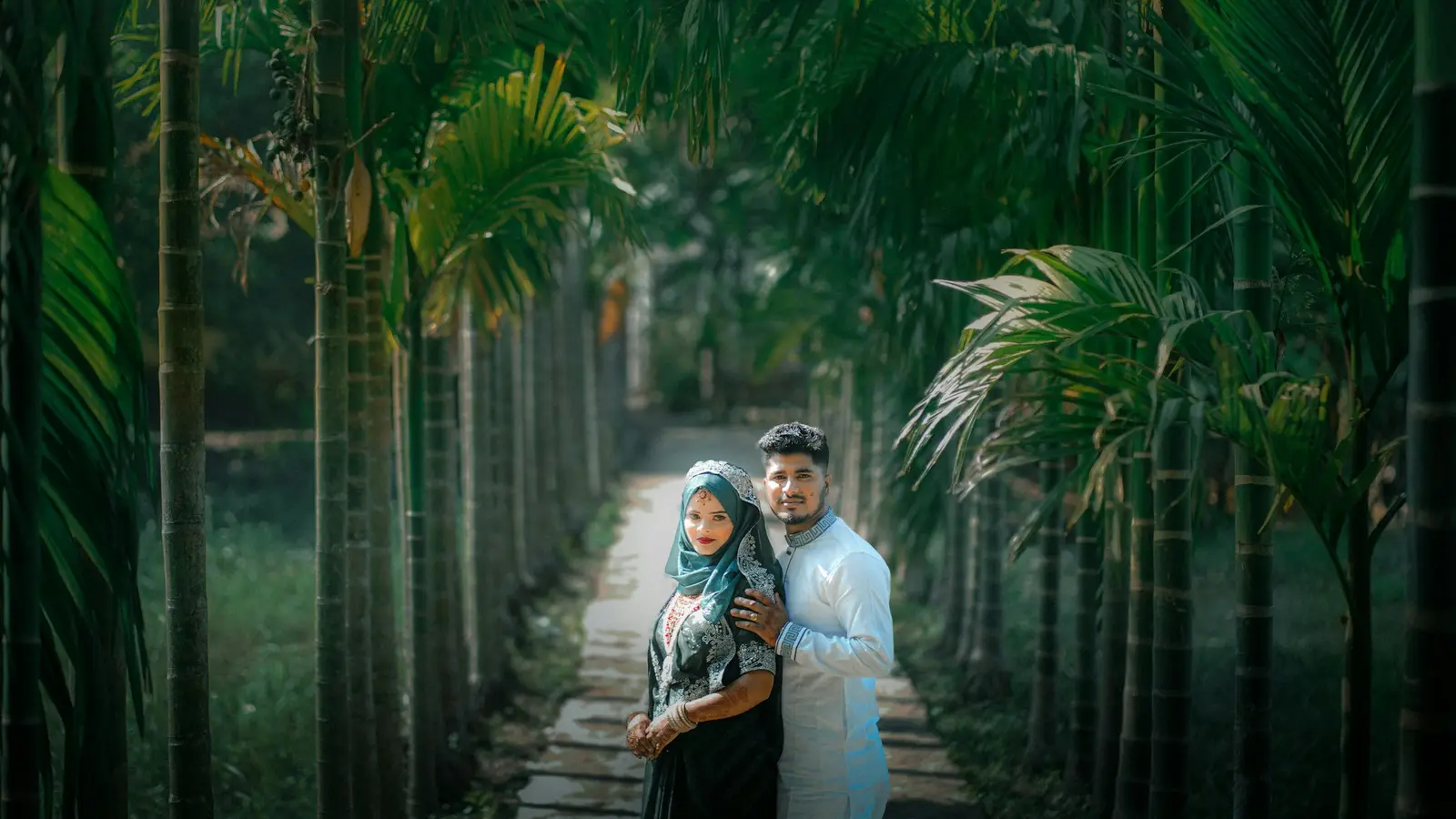 A newly married couple, the bride in a hijab, standing together on a path lined with palm trees.