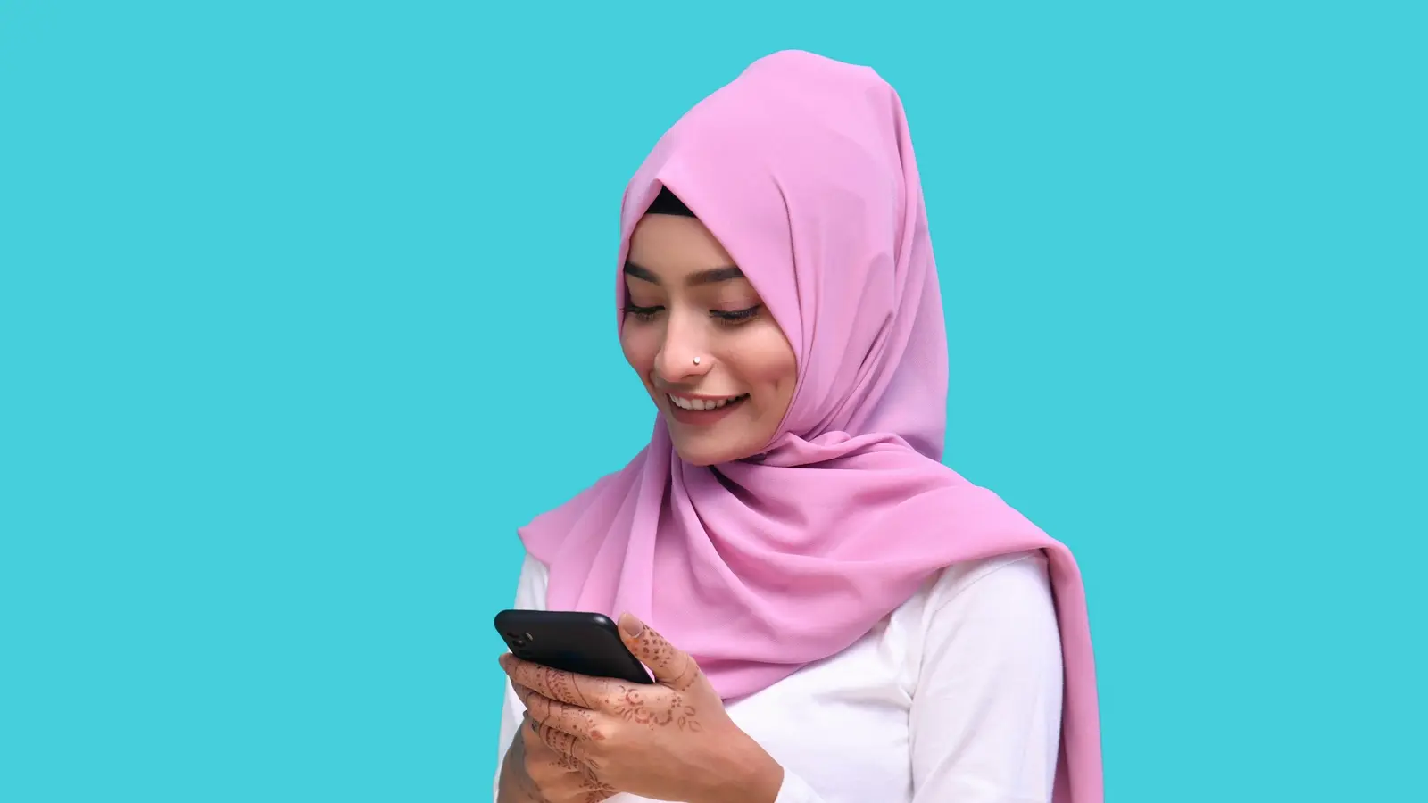 A smiling young woman in a pink hijab reading a message on her phone.