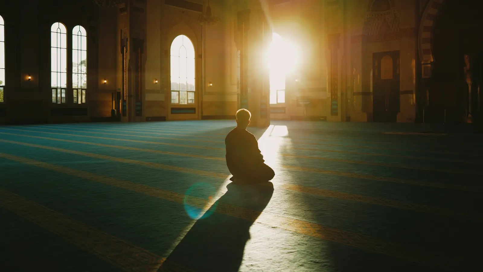 A man sitting alone on the carpet of a large mosque prayer hall as morning sunlight streams through the windows.