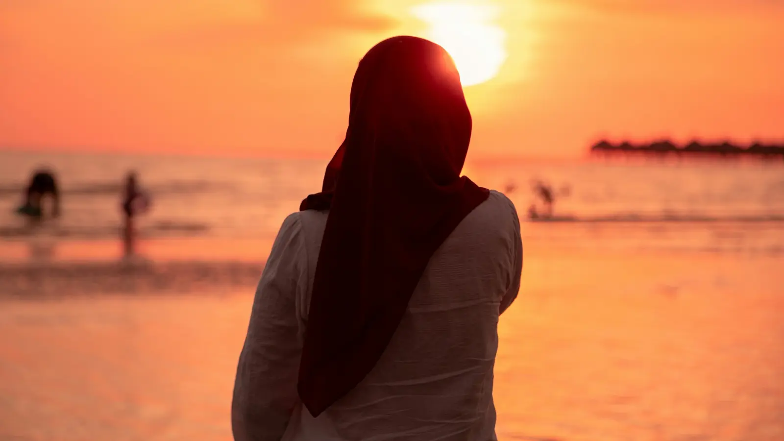 A woman in a dark hijab standing on a beach, looking out at the sun setting over the sea.