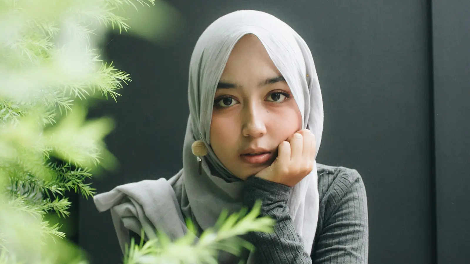 A young woman in a grey hijab resting her chin on her hand, deep in thought.