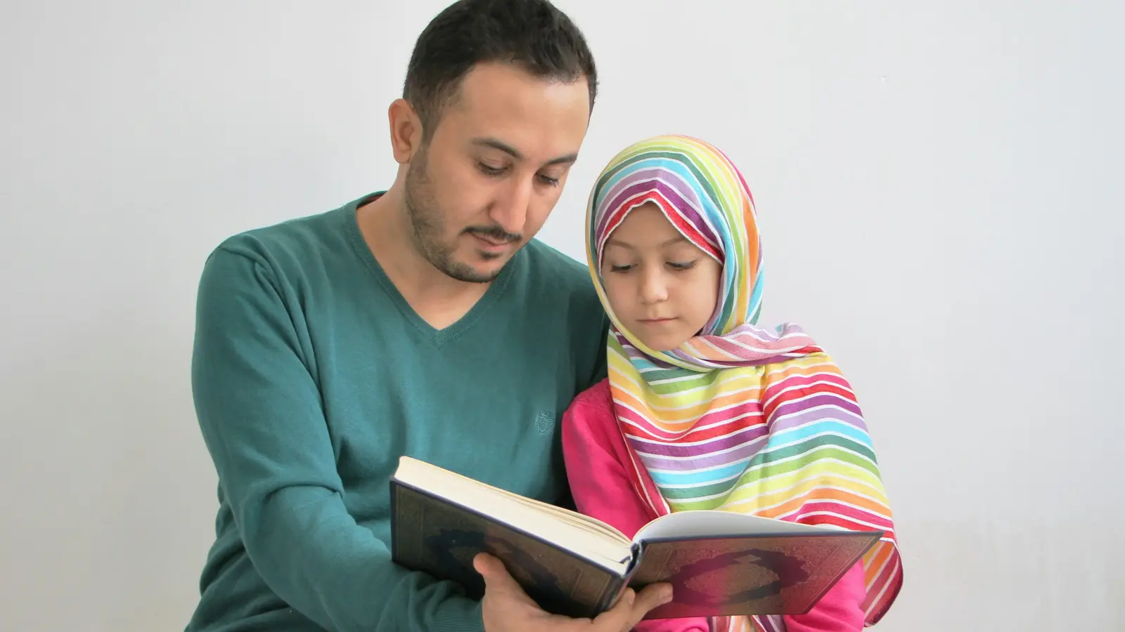 A father in a green jumper reading the Qur’an with his young daughter, who wears a colourful hijab.