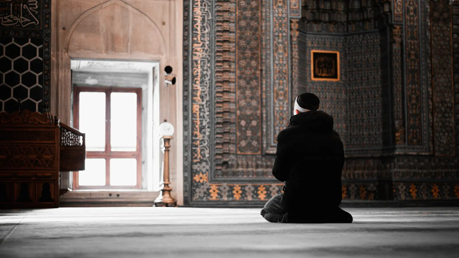 A man in a dark coat sitting on the floor of an ornate tiled mosque, facing a window full of light.